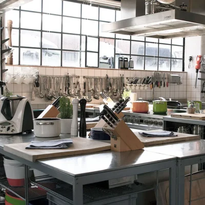 moderne-profikueche-edelstahl-arbeitsflaechen-kochutensilien Moderne Profiküche mit großen Fenstern, Edelstahl-Arbeitsflächen und Kochutensilien an der Wand