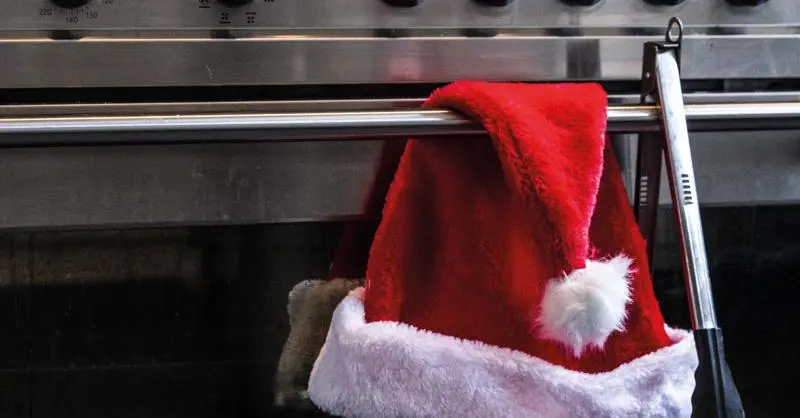Red Santa hat with white fur trim hangs on a stainless steel oven door handle in a kitchen kitchen scene.