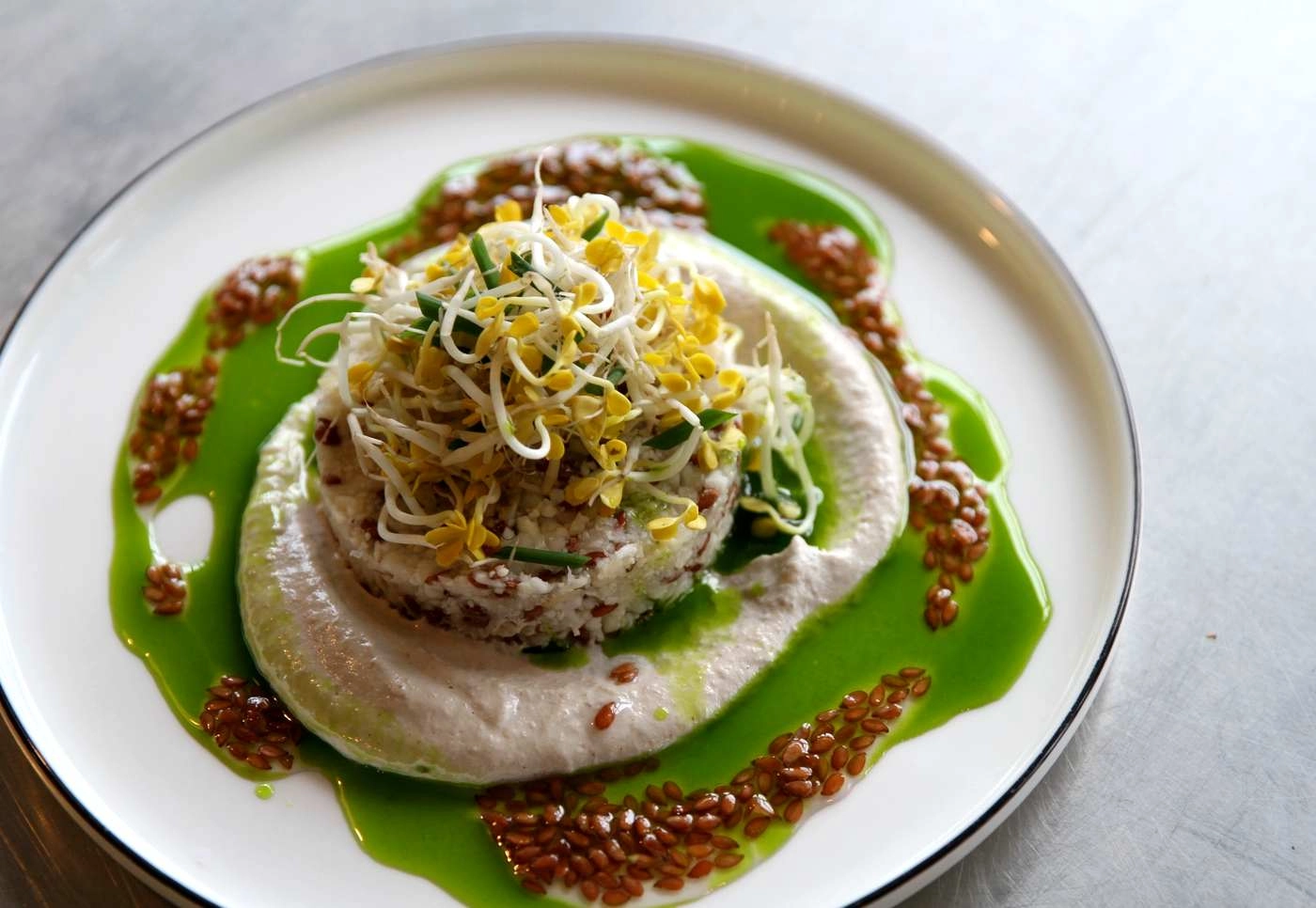 IMG_8881_web - KETAO | FRISCH. LECKER. ANDERS. Stacked grain patty topped with sprouts and microgreens, surrounded by bright green sauce and sesame-like seeds on a white plate (vegetarian main).