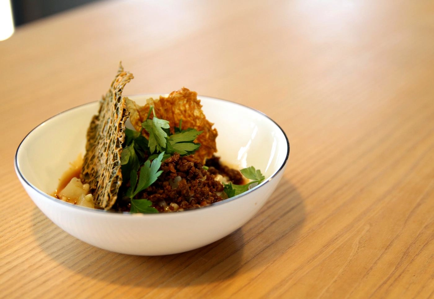 IMG_8897_web - KETAO | FRISCH. LECKER. ANDERS. White bowl on a wooden table containing a crispy lattice chip, fresh parsley, and seasoned crumbled meat with sauce, all arranged attractively as a plated dish.