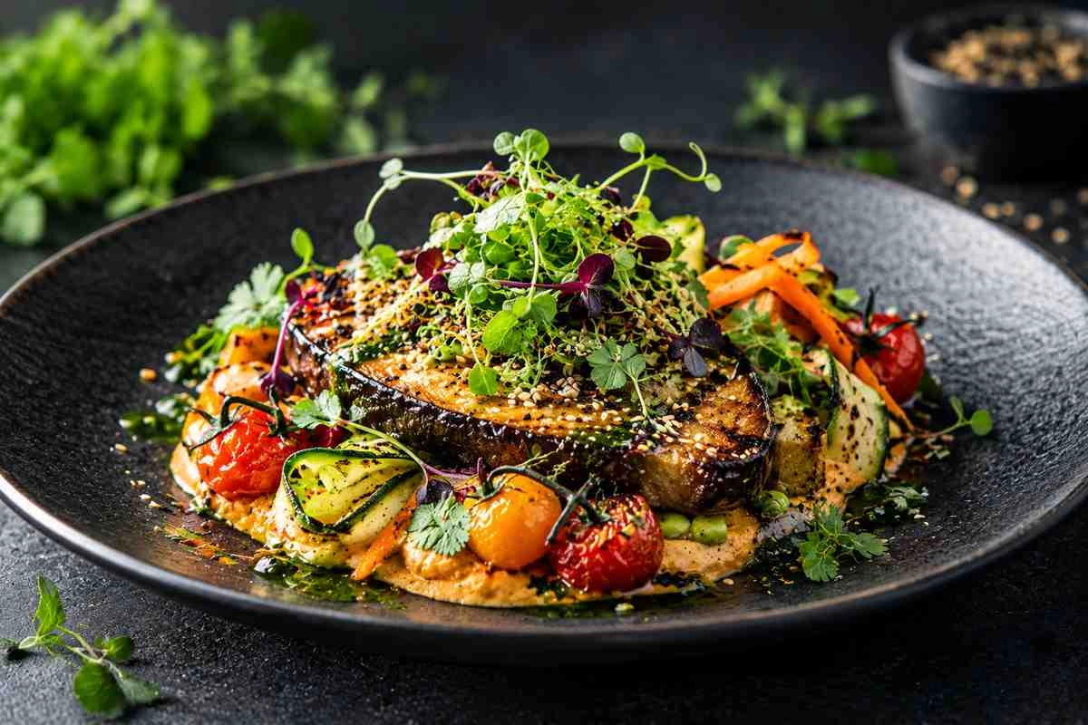 ketao_vegan_02 - KETAO | FRISCH. LECKER. ANDERS. Seared steak atop a bed of sauce with roasted cherry tomatoes, zucchini ribbons, and microgreens on a dark plate.